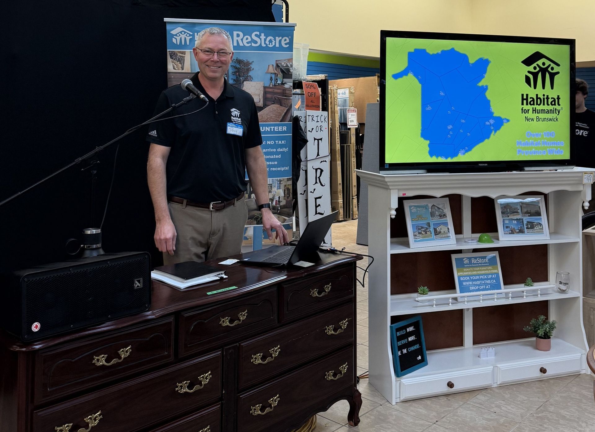 Perry standing beside a desk and display shelf, presenting a map on a screen in a booth.