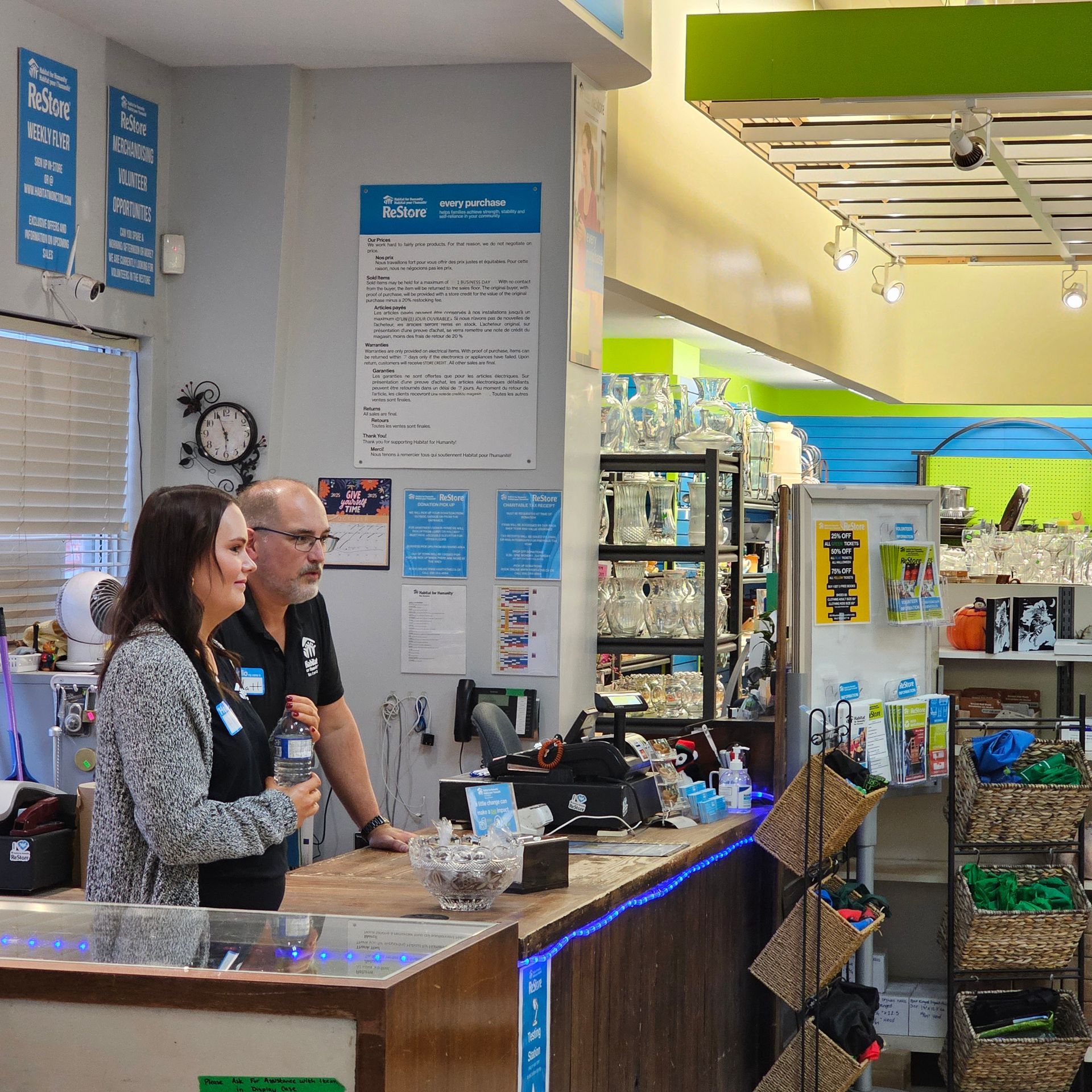 Two staff at a Restore counter with blue signage and shelves of packaged goods in the background