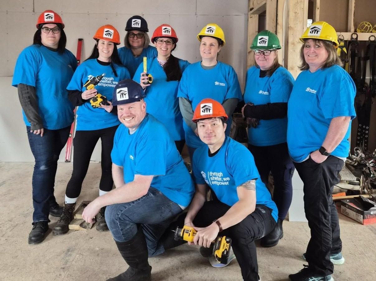 Group of Habitat New Brunswick volunteers in winter hats and masks posing outside a house, some holding paint cans.
