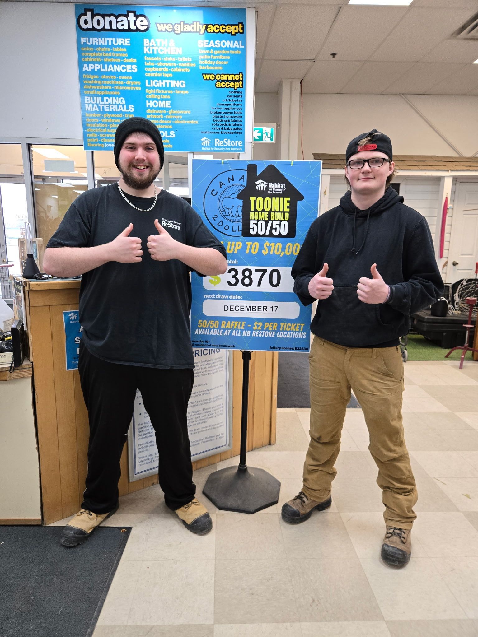 Two people standing indoors beside a blue Habitat New Brunswick donation sign, both smiling and giving thumbs up.