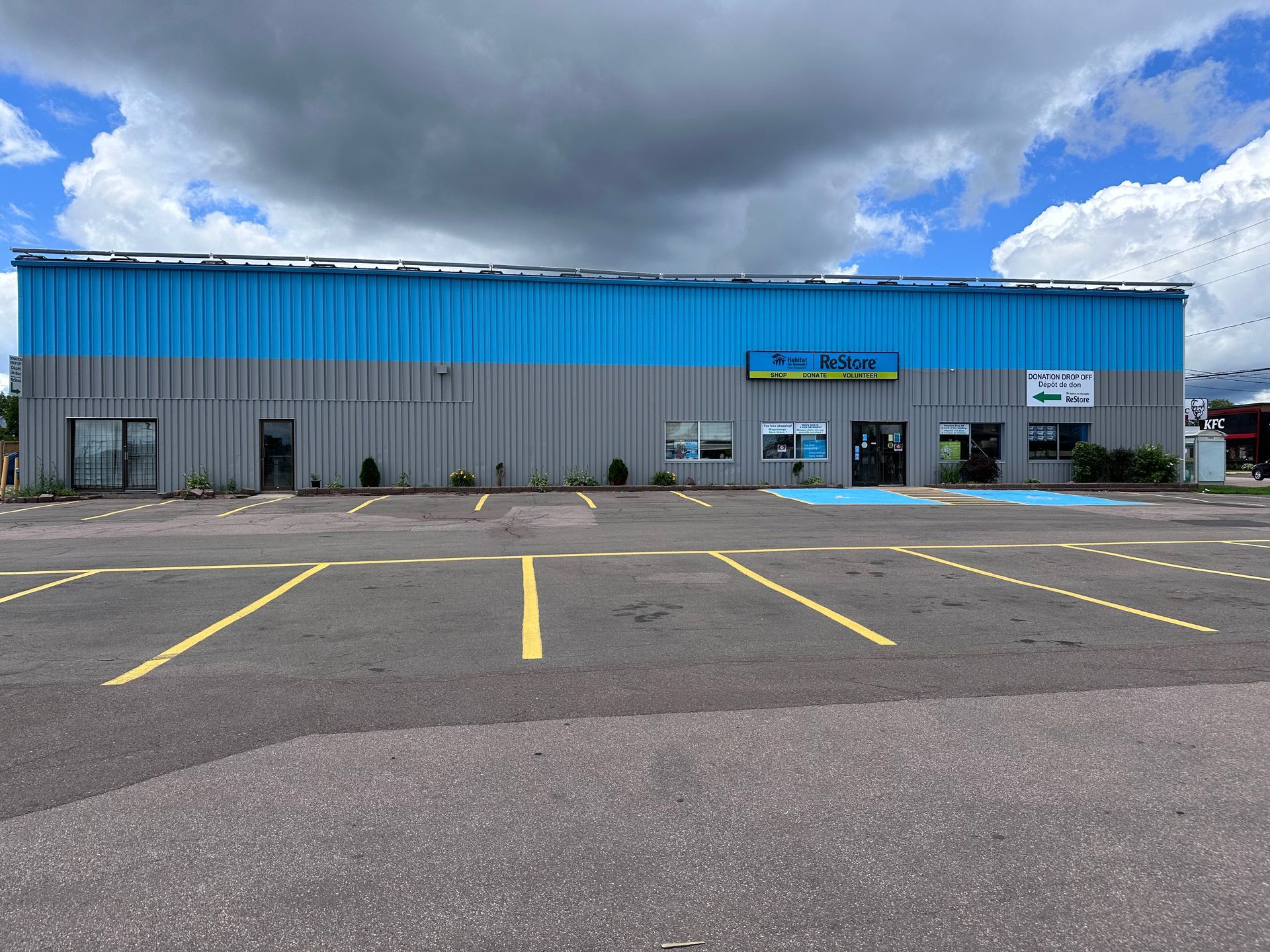 Moncton Restore, Empty parking lot in front of a gray building with a bright blue roof under cloudy skies