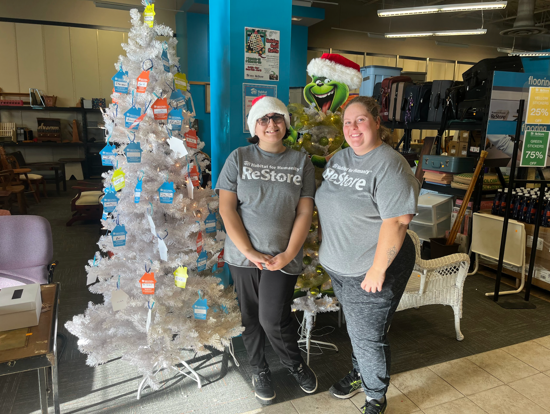 Two women stand beside a decorated silver Christmas tree in a store. Habitat New Brunswick volunteers.