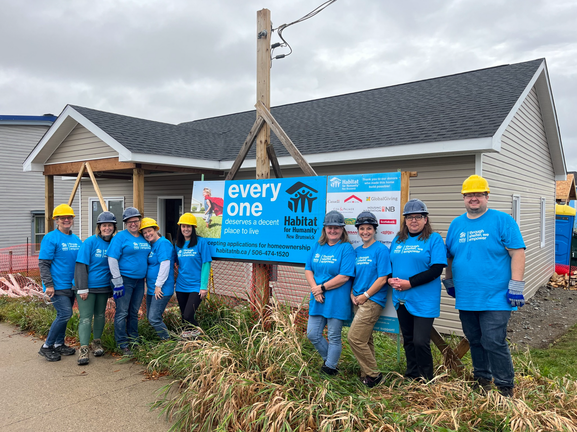 Habitat New Brunswick volunteers in blue shirts and hard hats stand with a “every one” sign in front of a house