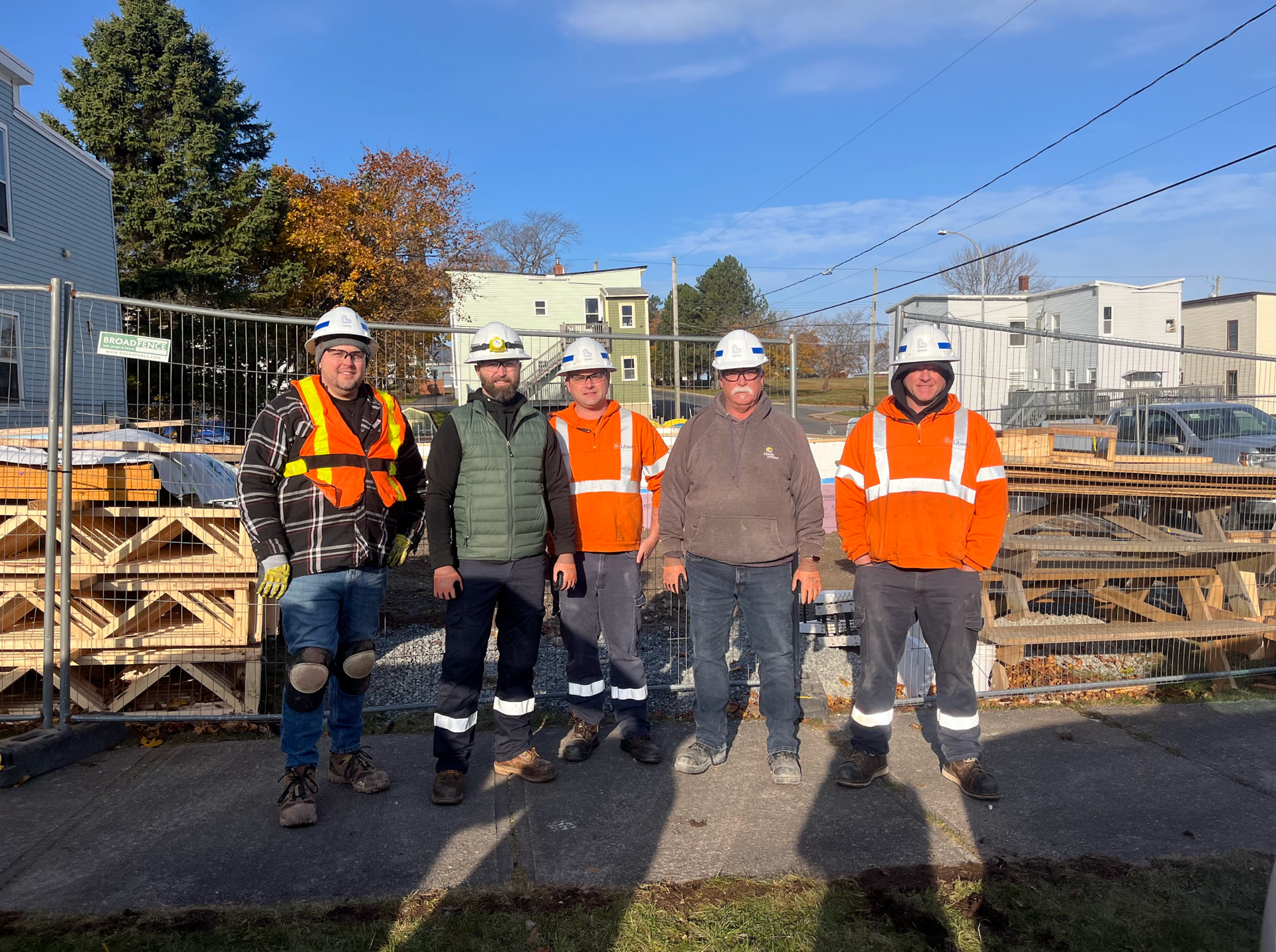 Five workers in hard hats and safety vests standing outdoors by stacked wooden pallets, Habitat New Brunswick volunteers