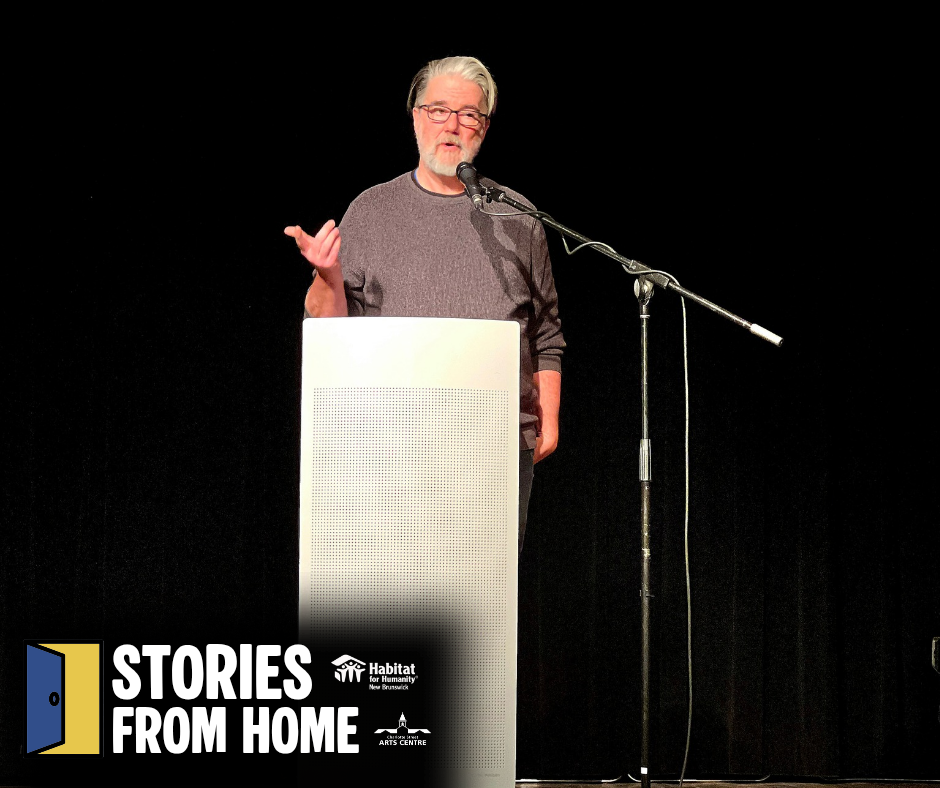 Habitat New Brunswick, Man speaking at a podium on a dark stage with a microphone and “Stories From Home” sign.