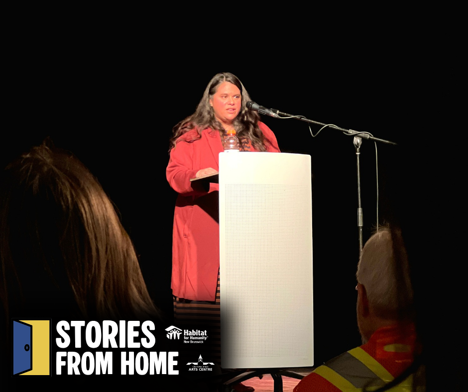 Woman in a red dress speaking at a podium on stage Habitat New Brunswick, with a microphone and audience silhouettes.