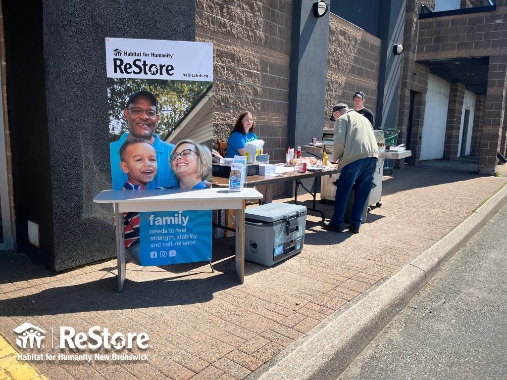 Habitat New Brunswick volunteers staffing a ReStore table outside a building, with sign, flyers, and donations bin