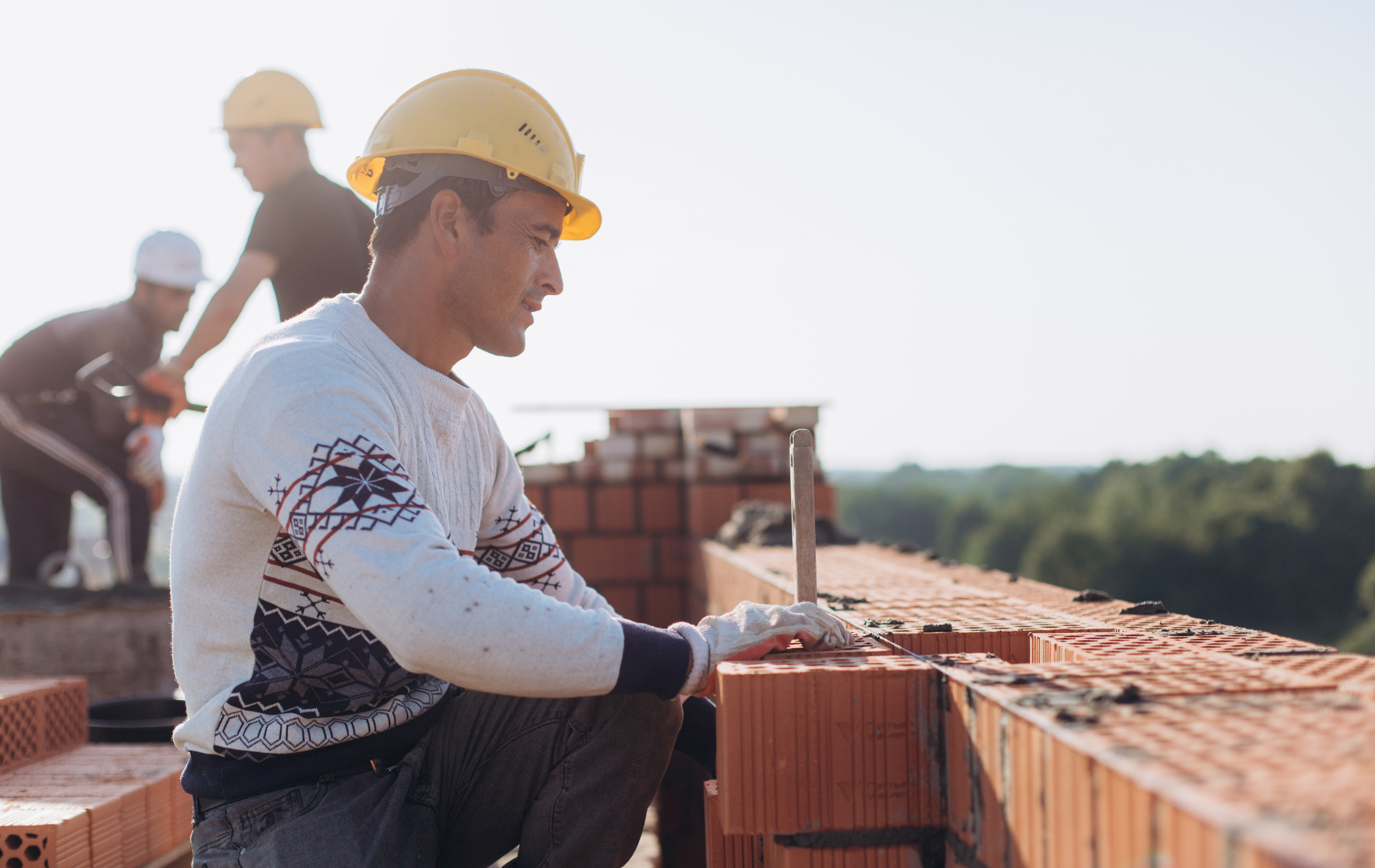 Construction worker in a yellow hard hat laying bricks on a rooftop with others.