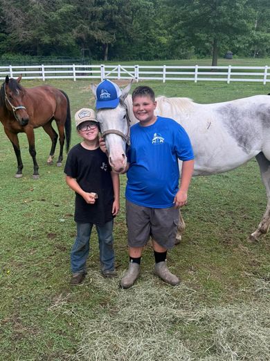 Two boys pose with a white horse and two other horses in a grassy paddock with a white fence.