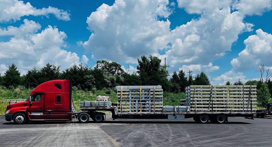 Red semi-truck hauling stacked pallets on a flatbed trailer under a cloudy blue sky.
