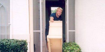 Man carrying a box through a doorway, with a screen door and bushes in front of a white building.