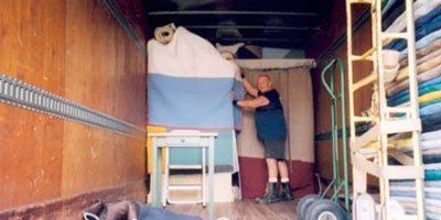 Man loading furniture in a moving truck. Light-colored cabinet, tall wrapped items, and hand truck visible.
