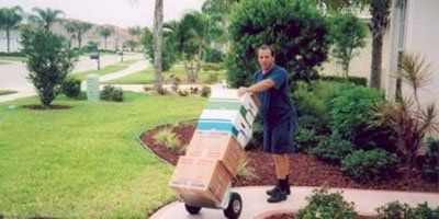 Man pushing a hand truck loaded with moving boxes on a sidewalk in front of a house.
