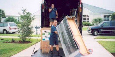 Movers unloading furniture from truck in front of a house. One man carrying a wrapped item, the other in the truck.
