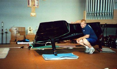 Man squatted near grand piano on stage in a room with organ pipes, cross, and two tall poles.