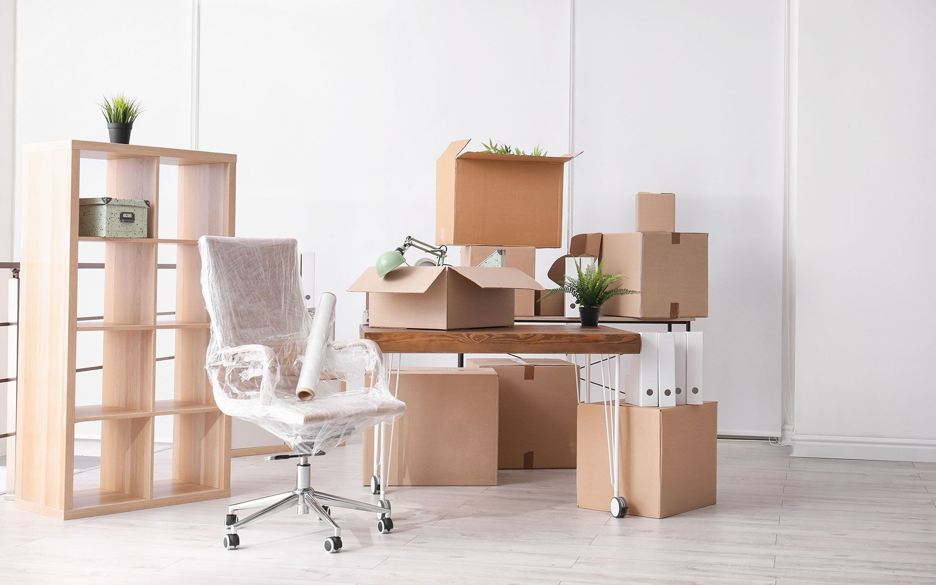 Office space being prepared for a move; packed cardboard boxes and wrapped chair surround a desk and shelves.