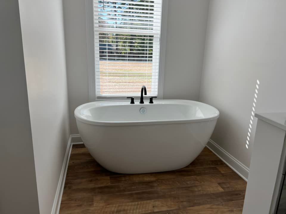 Freestanding white bathtub in a bathroom, under a window with blinds, brown floor, black faucet.