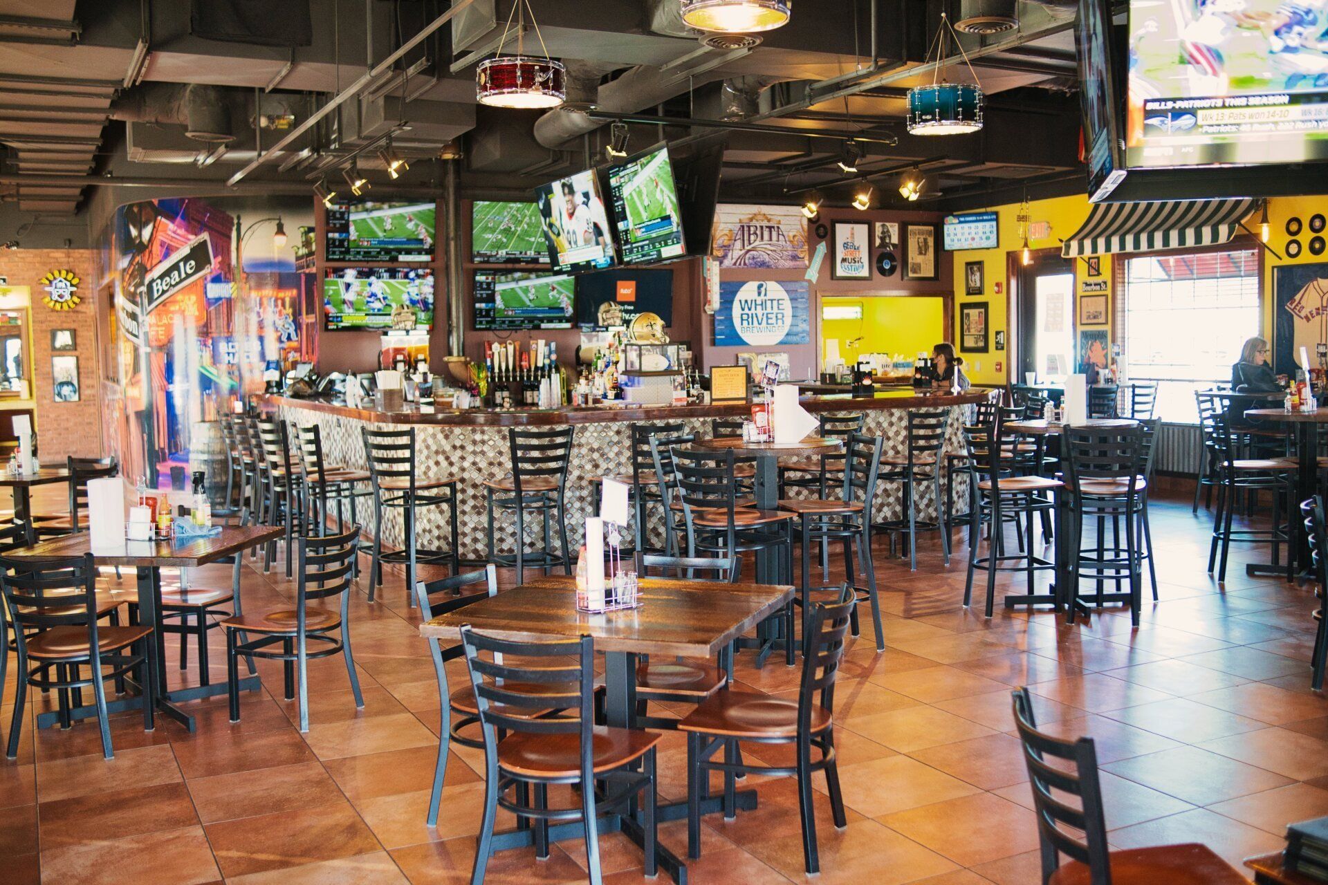 Empty sports bar with tables, chairs, TVs, and a polished wood floor under colorful hanging lights.