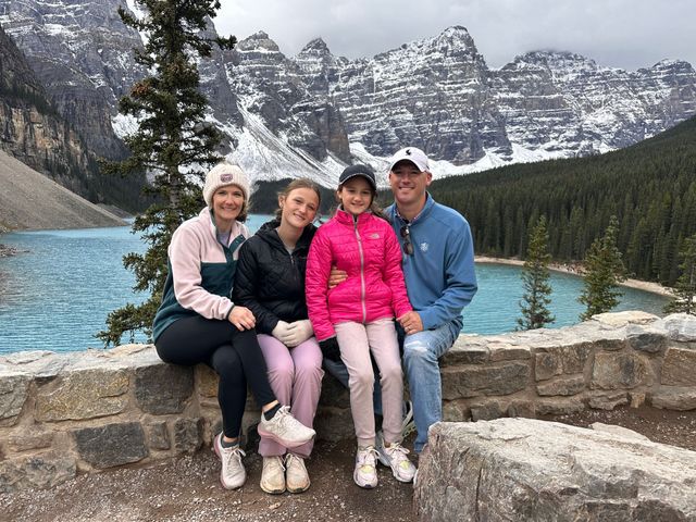 Four people sitting on a stone wall by a turquoise lake with snowy mountains behind them