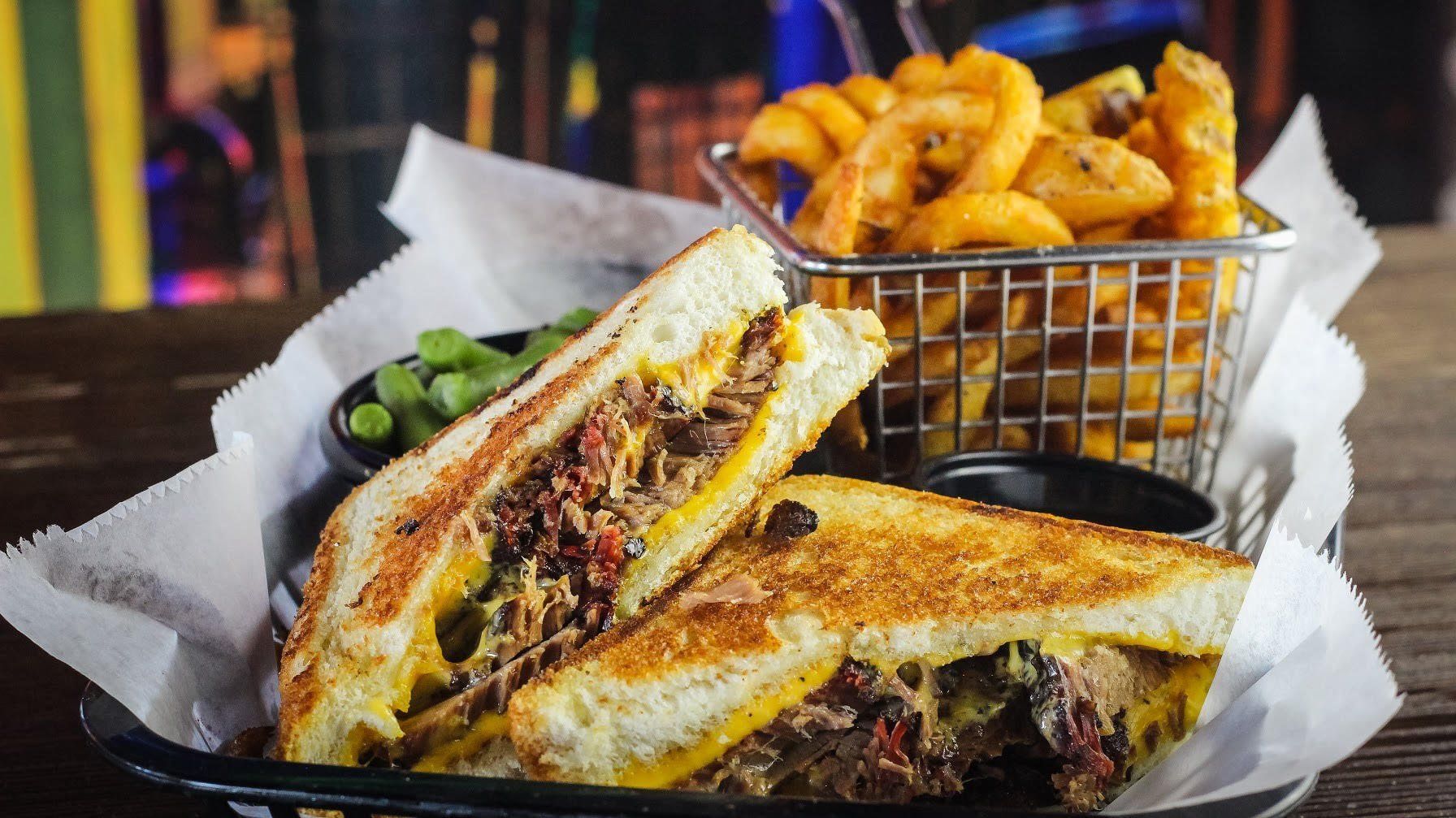 Grilled sandwich with melted cheese, fries in a metal basket, and pickles on a tray.
