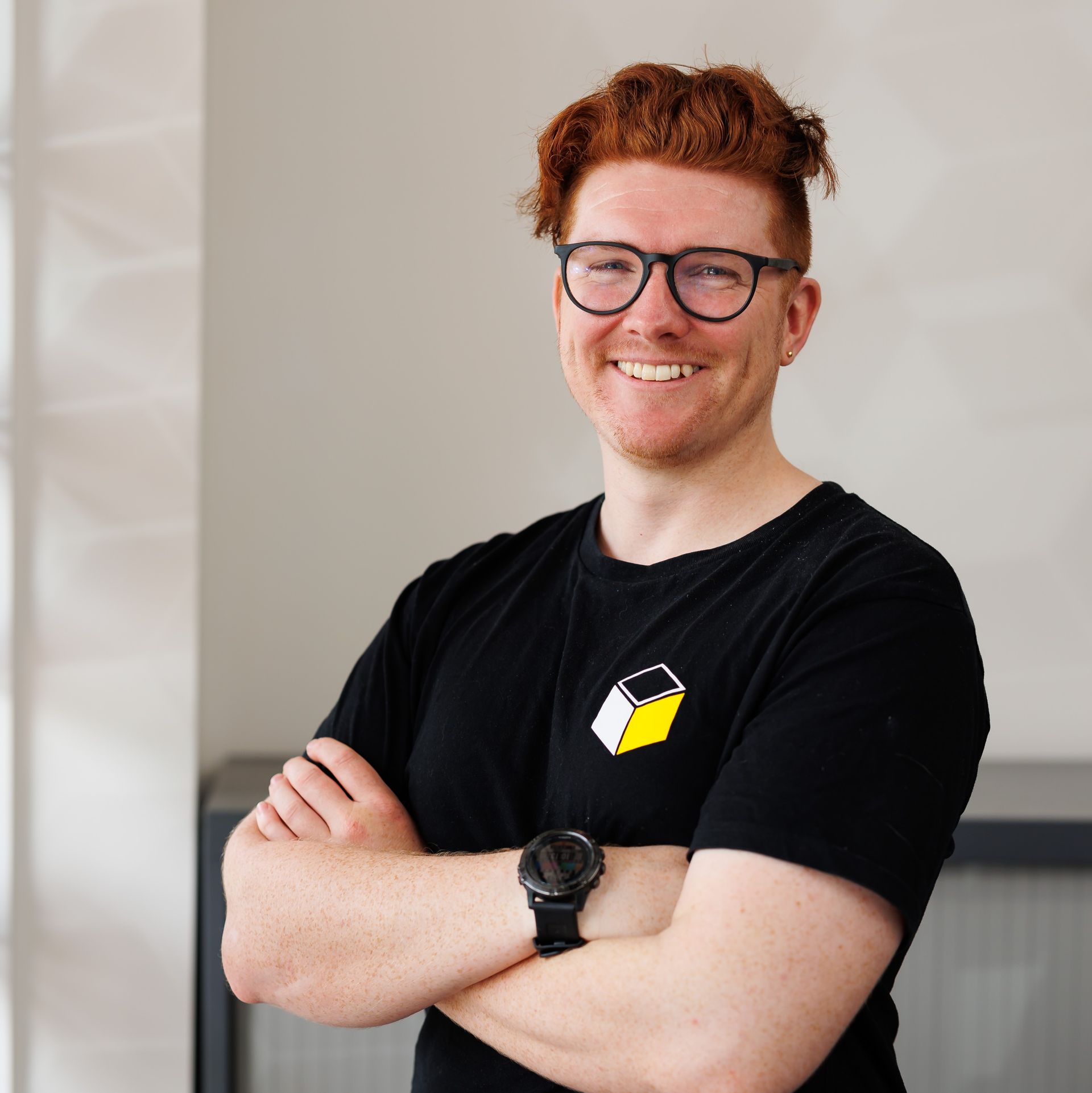 A man wearing glasses and a black shirt is smiling with his arms crossed