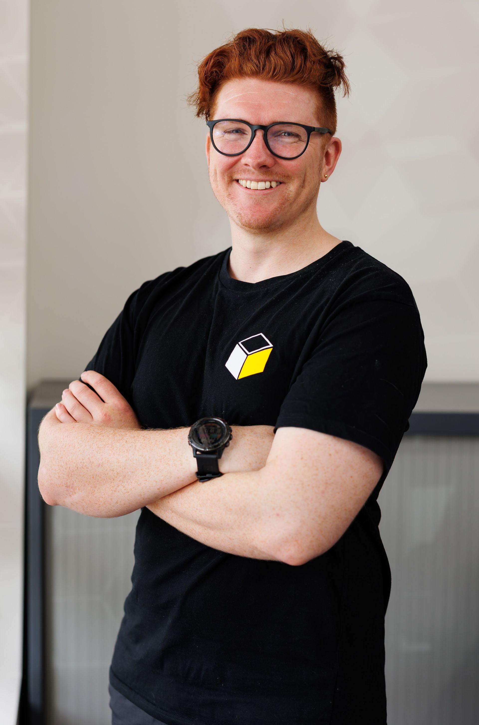 A man wearing glasses and a black shirt is standing with his arms crossed and smiling.