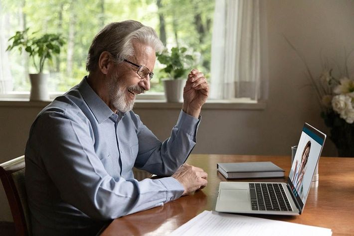 Man using a laptop at a wooden table, smiling during a video call in a bright home office