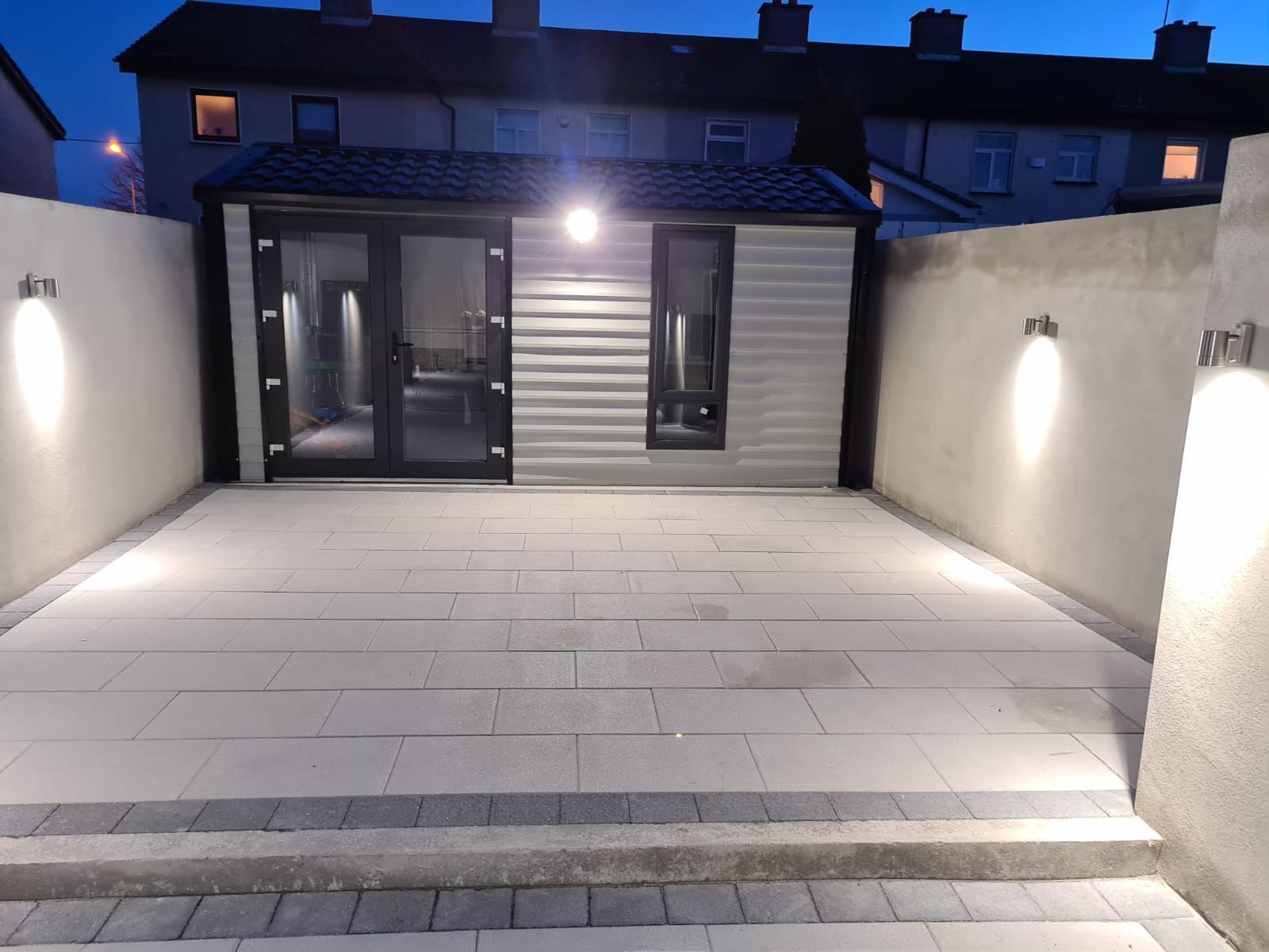 Backyard patio with lit walls and shed. Grey paving, walls, and exterior lights.