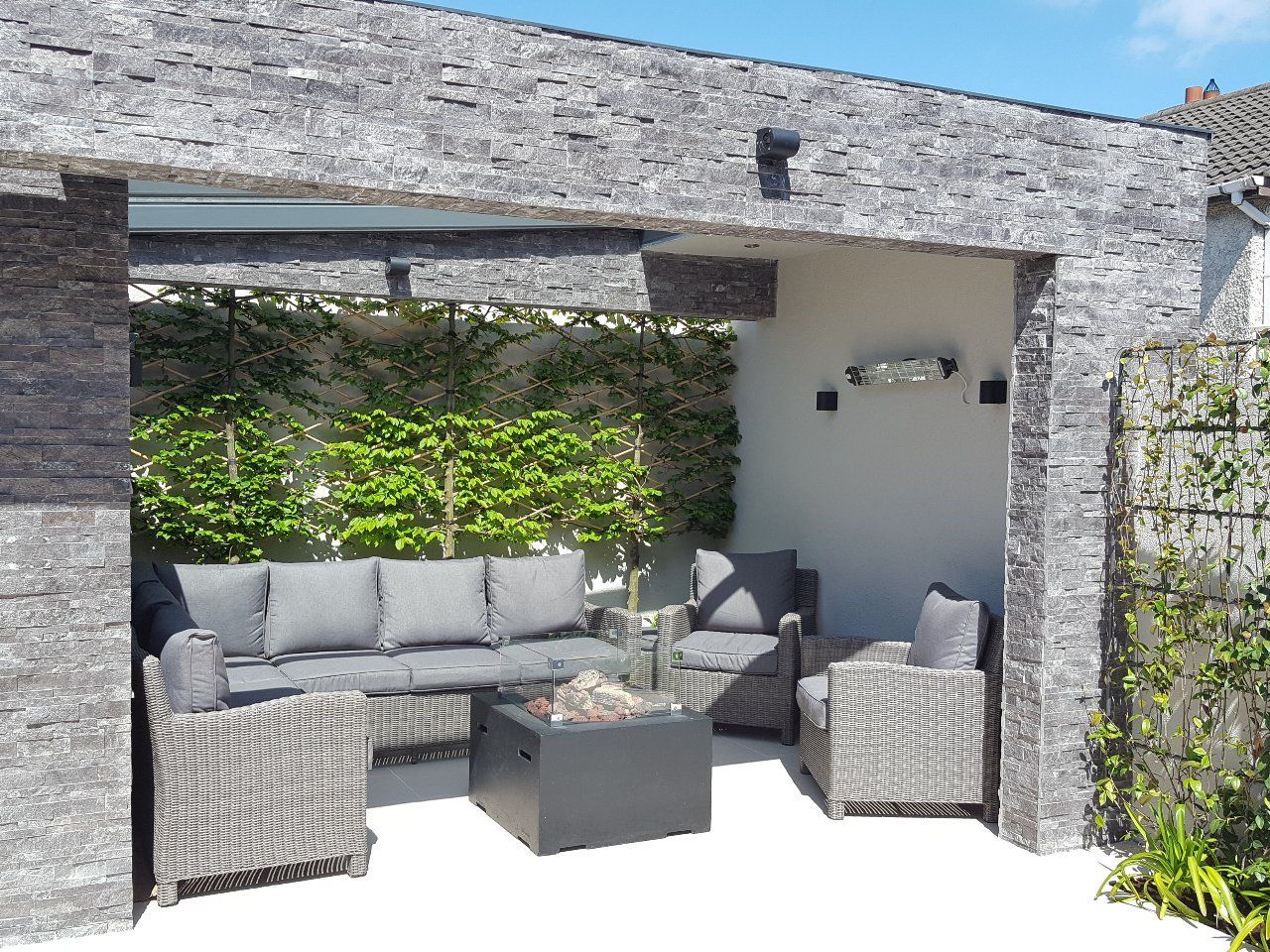 Outdoor seating area with gray stone walls, sofa, armchairs, and fire pit. Green foliage and a white patio.