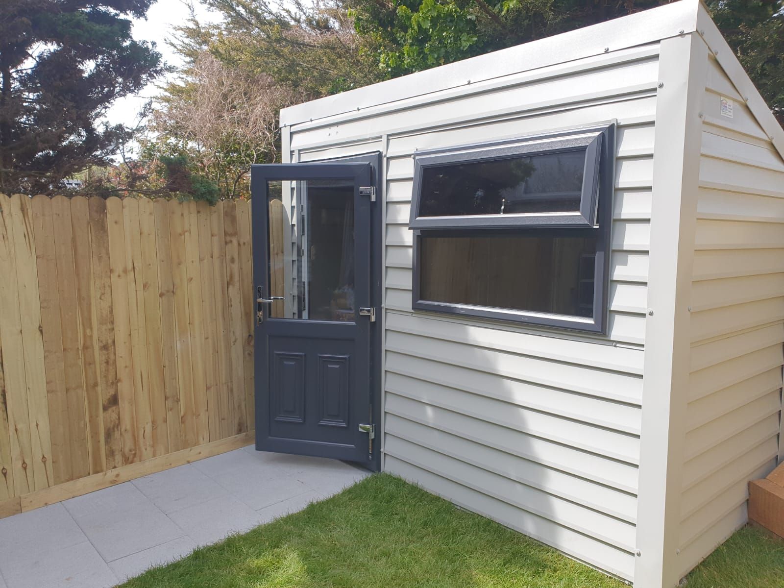 Small gray shed with an open door and window, set beside a wooden fence and grass.