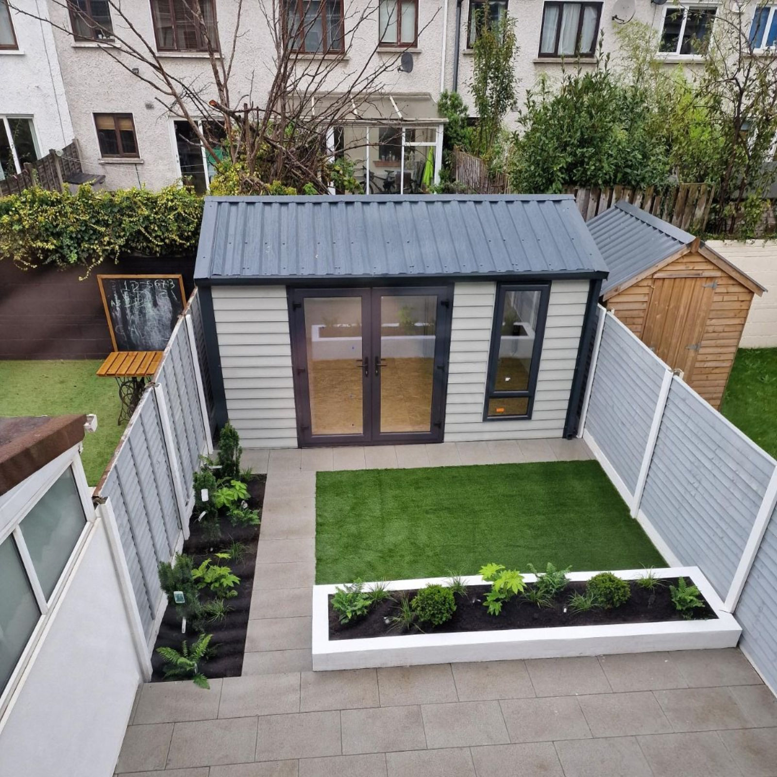 Backyard with a gray-sided shed, small lawn, flowerbeds, and a matching fence, next to a larger building.