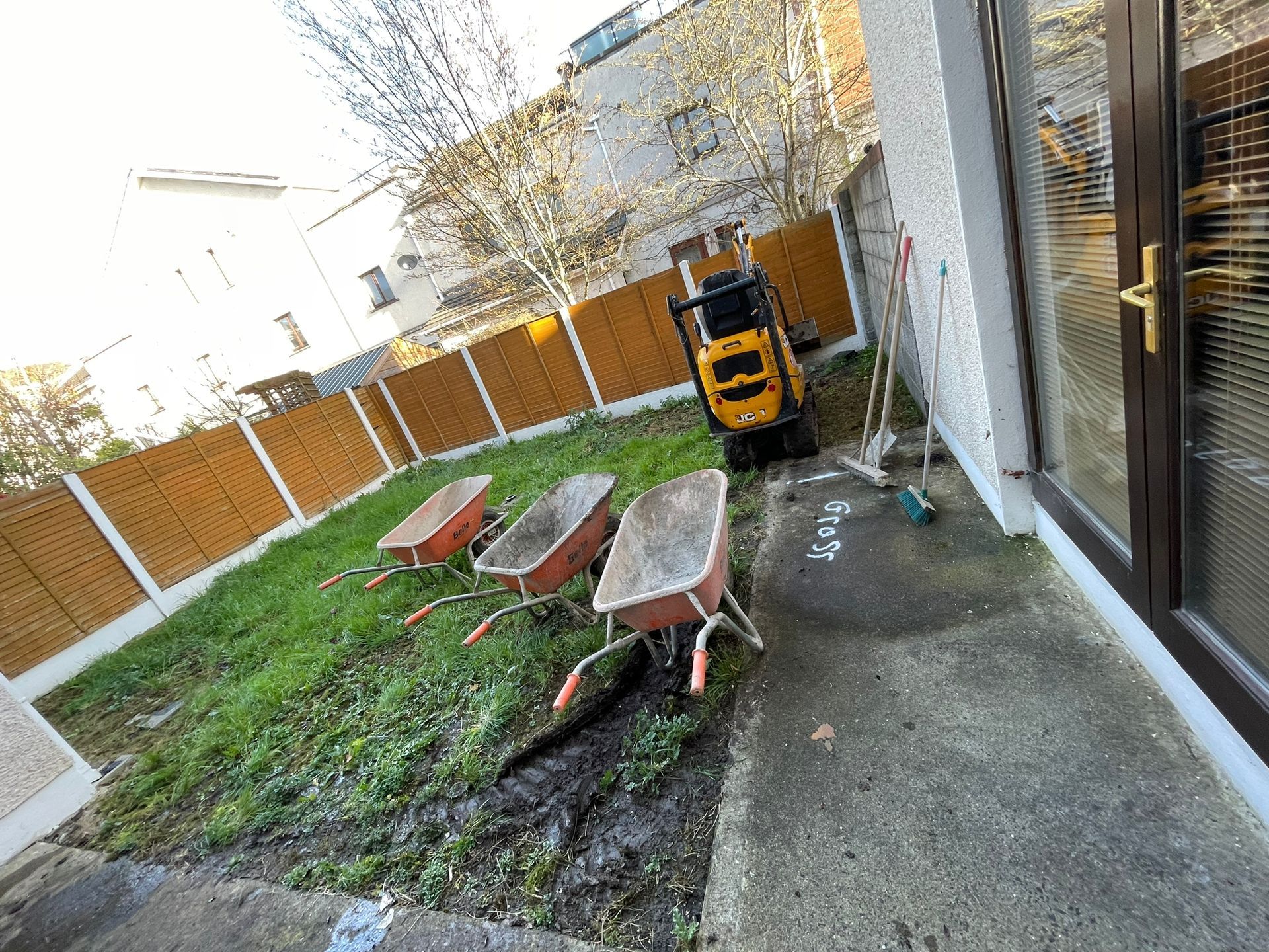 Backyard with wheelbarrows, small digger, and fence.  Concrete patio next to a door.  Grassy area.