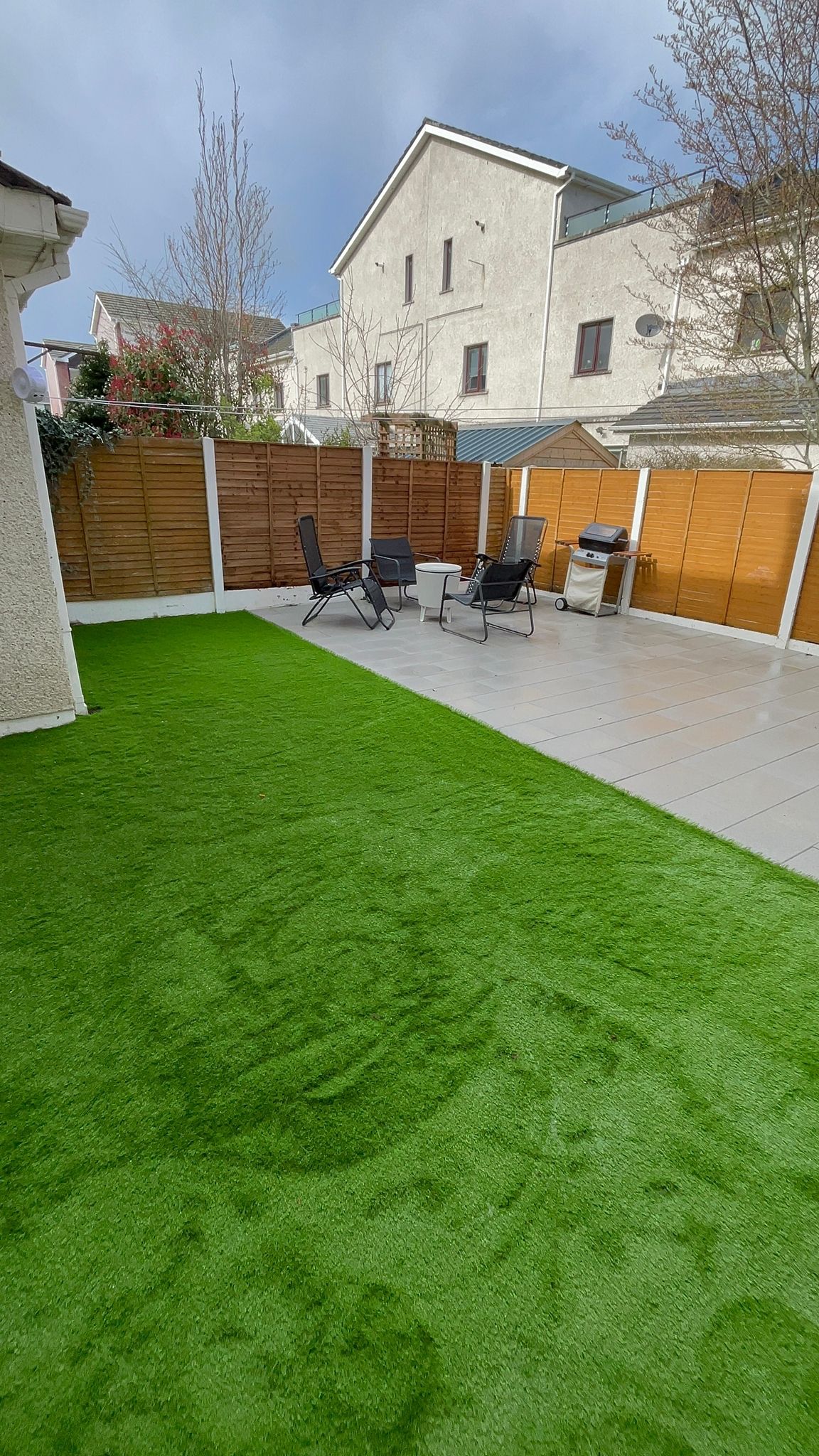 Backyard with green turf, patio, wooden fence, and seating. Buildings in background.