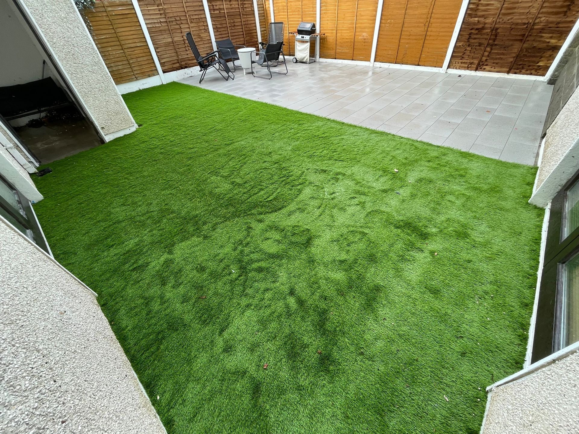 Small backyard with artificial turf and patio. Two chairs and a grill are on the patio. Wooden fence backdrop.