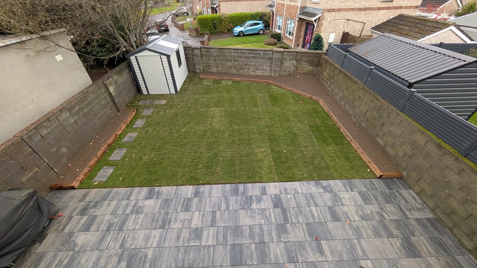 A backyard with a tiled patio, green lawn, small shed, and brick borders, surrounded by stone walls.