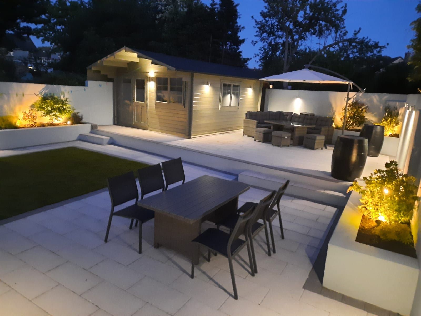 Outdoor patio with seating areas, a shed, and trees at dusk, illuminated with warm lighting.