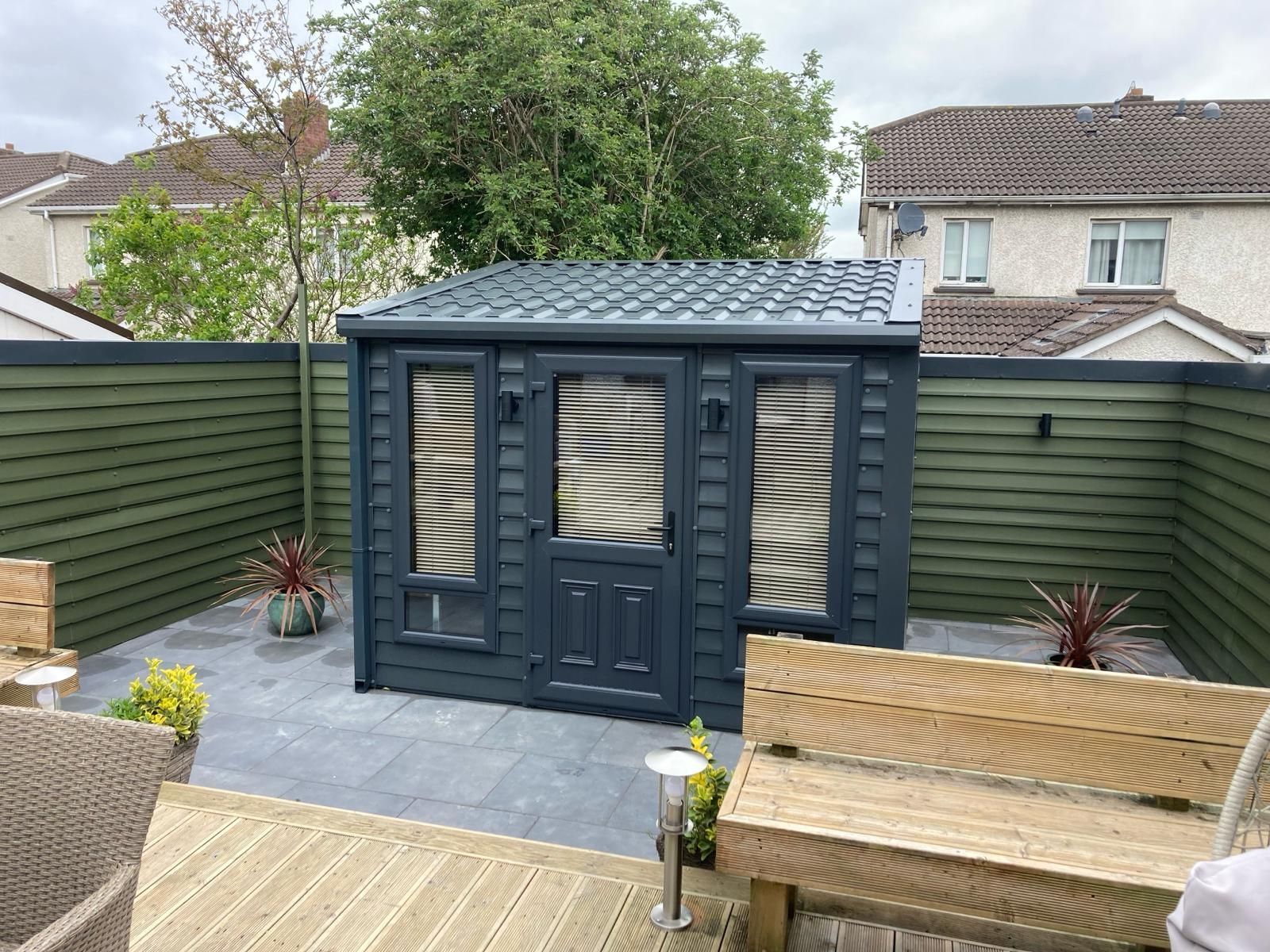 A dark gray shed with slatted windows and a matching fence, surrounded by a paved patio and seating.