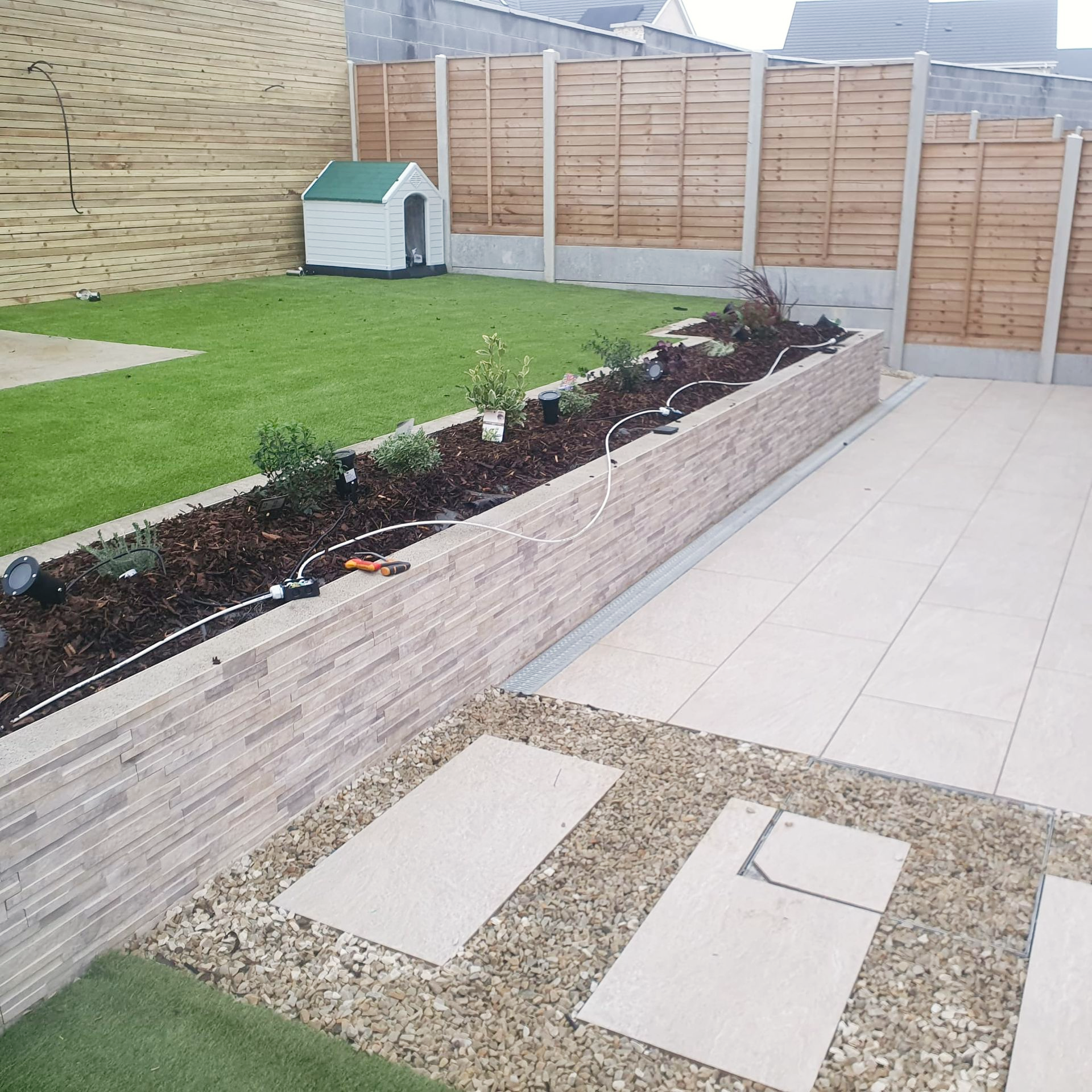 Backyard with a raised garden bed, lawn, stone patio, wooden fence, and dog house.