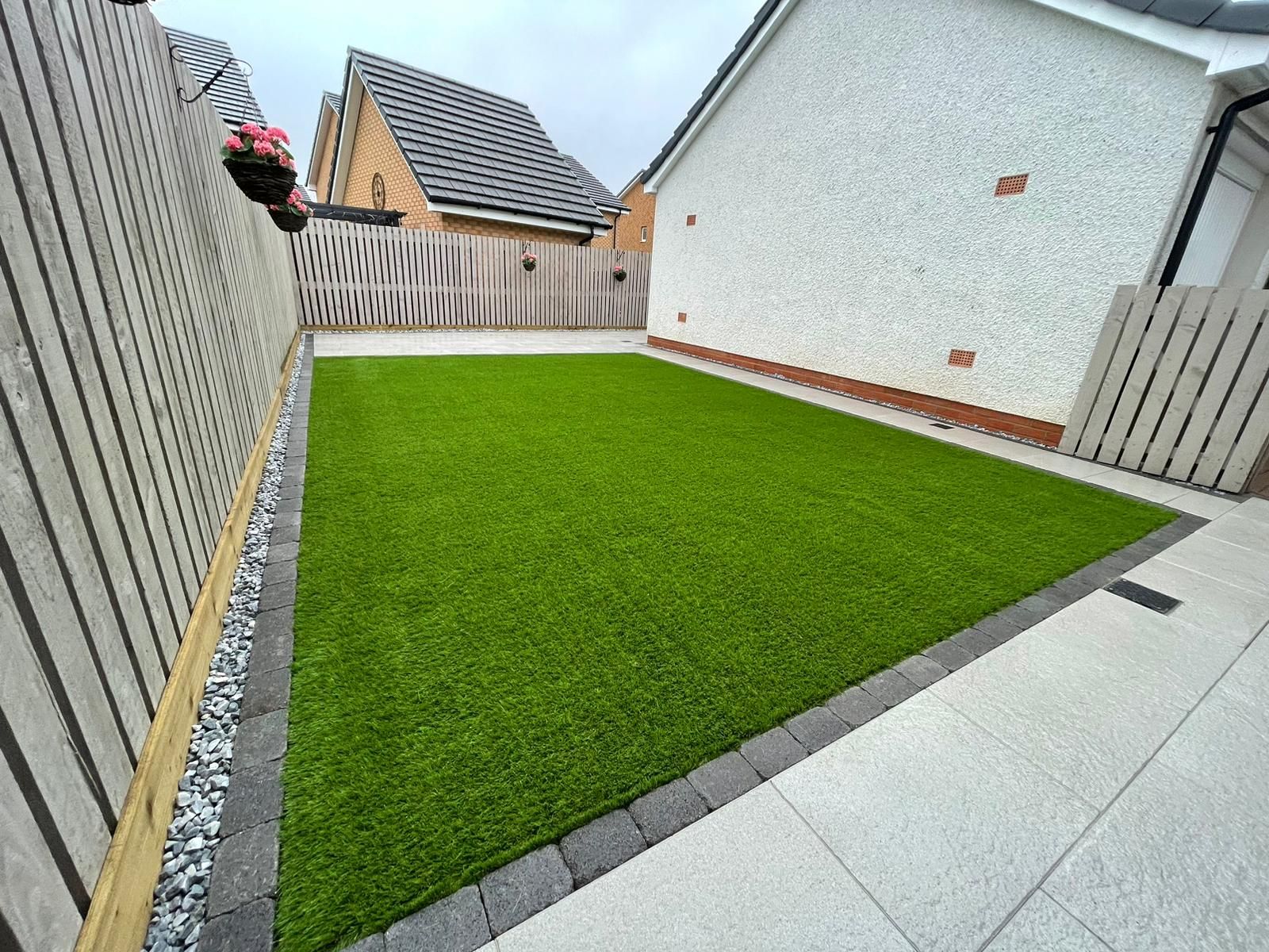 Green artificial lawn bordered by dark gray bricks and light paving stones, adjacent to a fence and house.