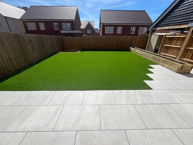 Backyard with green turf lawn, light gray patio stones, and a wooden fence. Houses in the background under a blue sky.