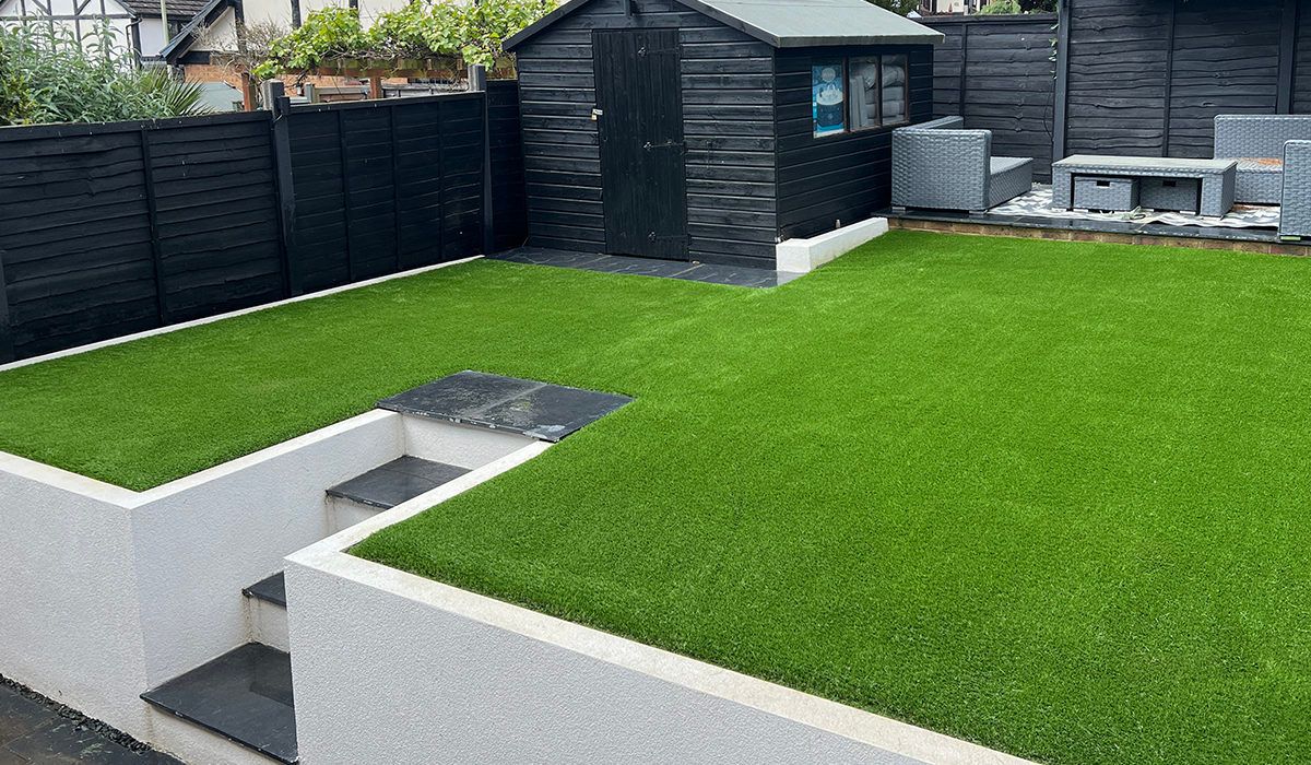 A backyard with artificial green grass, two raised levels, black shed and fence, and outdoor furniture.