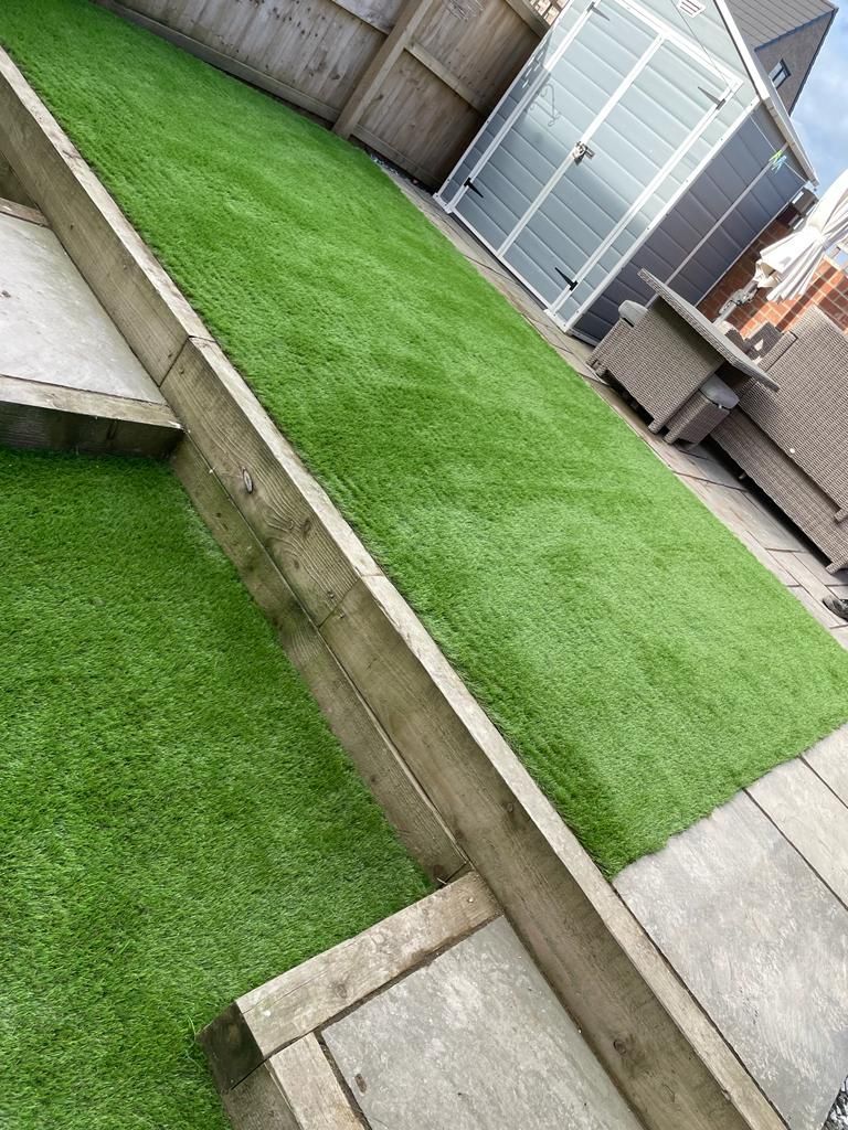 Green artificial turf in a sloped backyard, bordered by wooden beams and paving stones.
