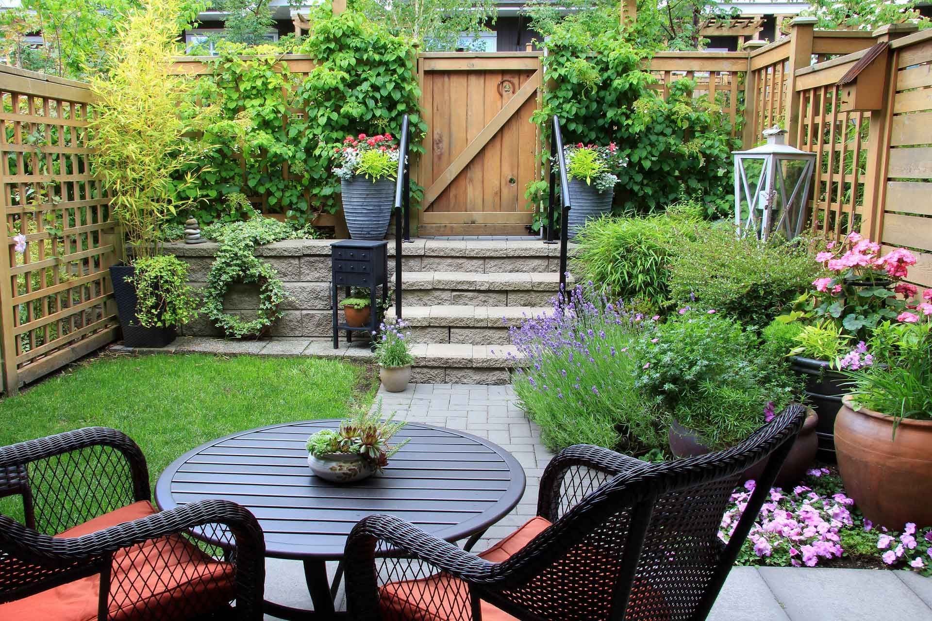 Cozy backyard patio with a round table and chairs surrounded by lush greenery, flowers, and a wooden gate.