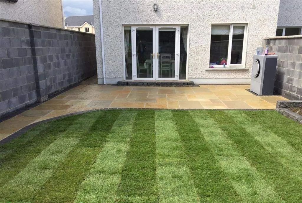 Backyard with striped green lawn, paved patio, and a light-colored house with glass doors.