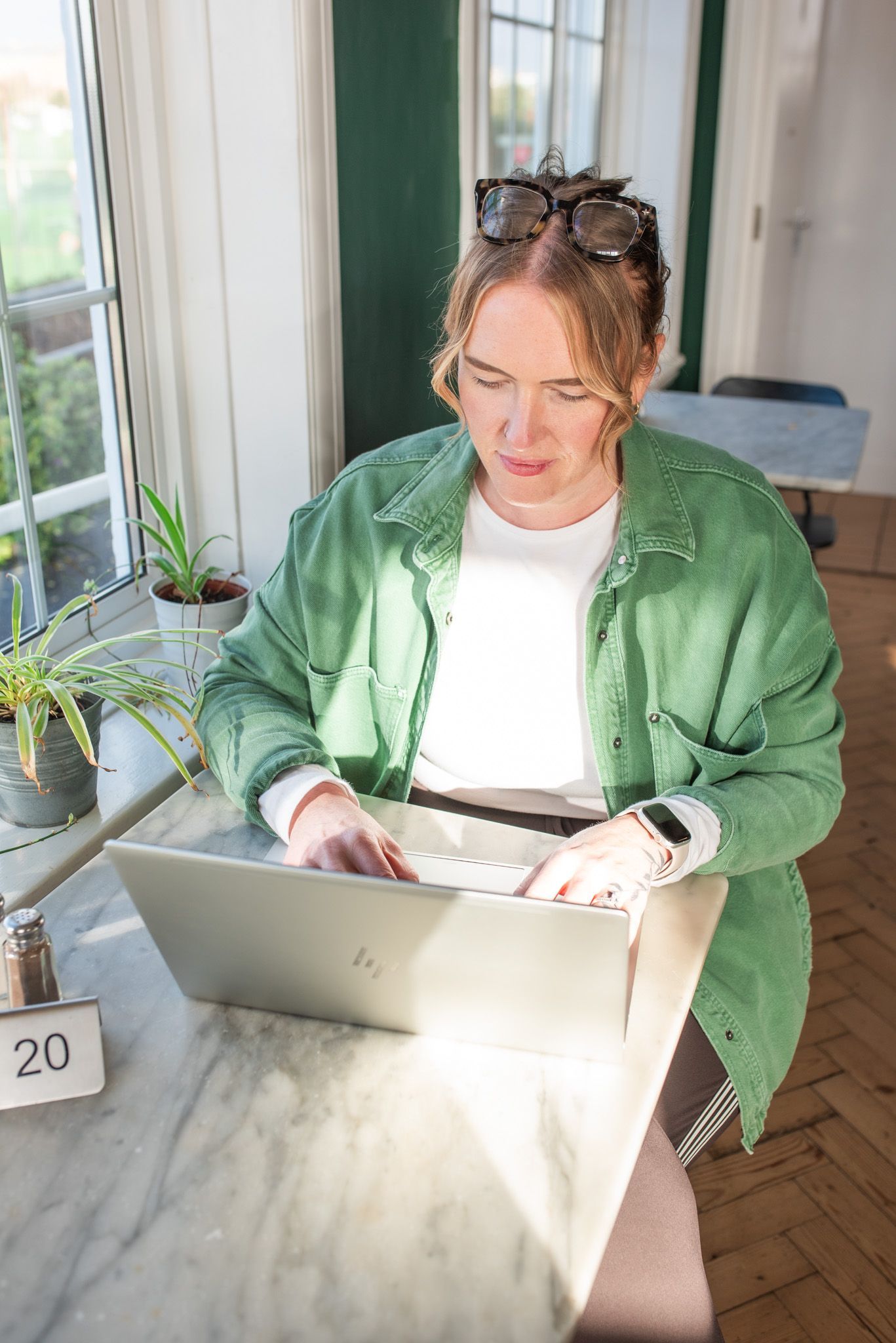 Person in green shirt working on a laptop at a table by a window, sunglasses on head.