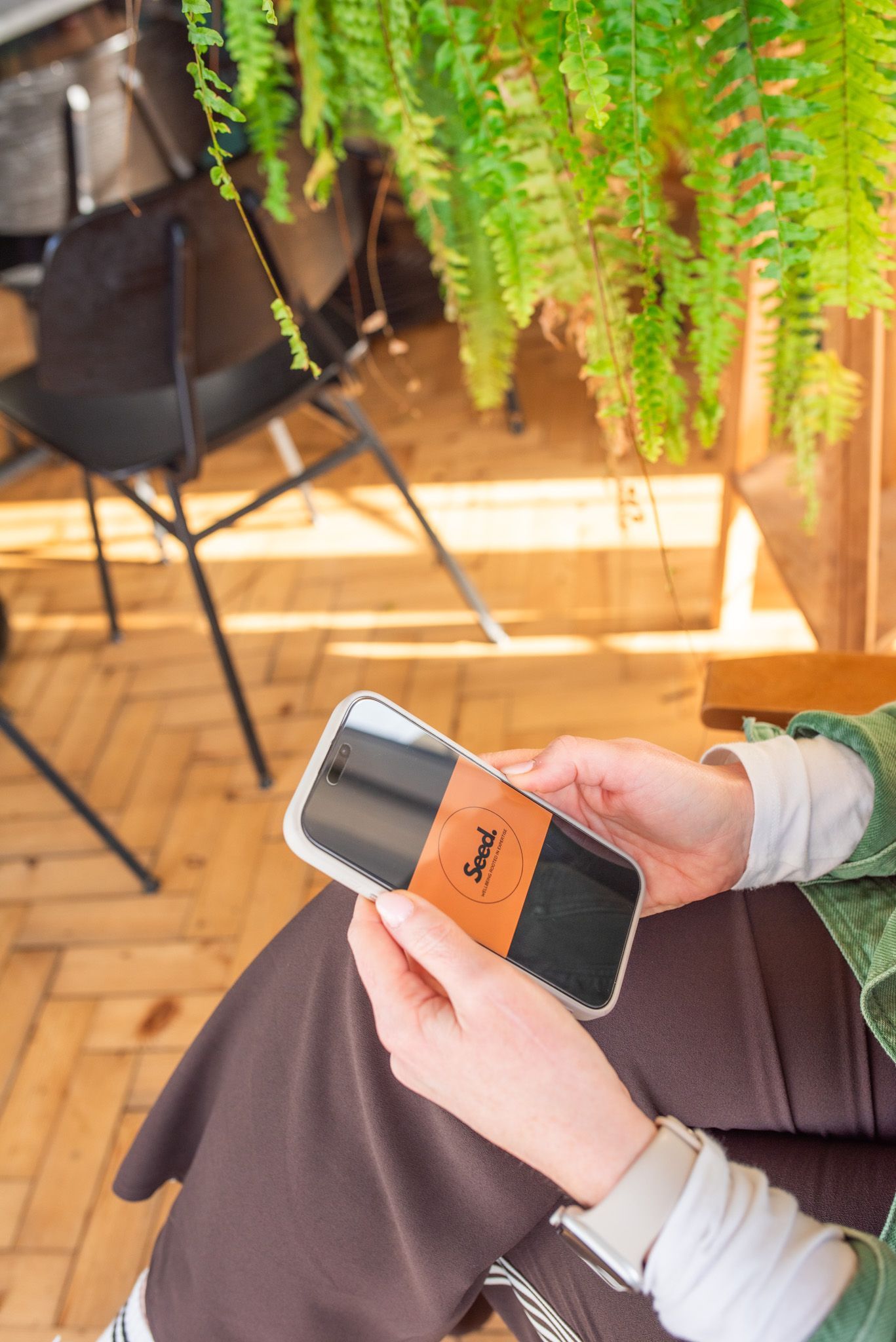 Person holding a phone with a brown label on the screen, seated outdoors. Ferns hang above.