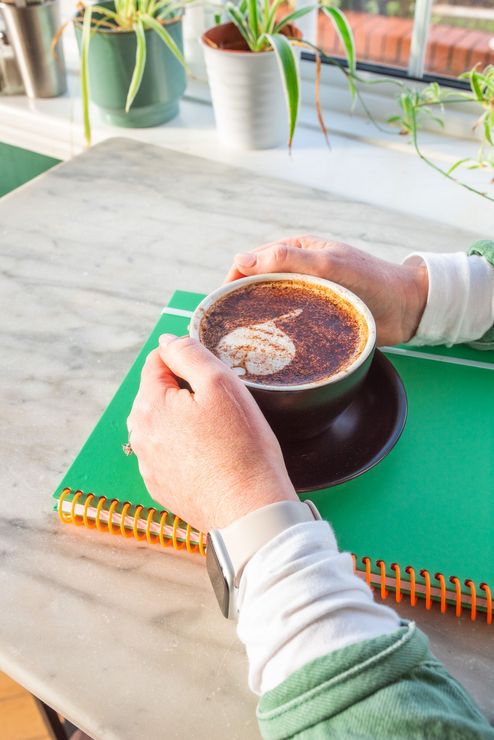 Hands holding a coffee cup on a green notebook, sunlight on a marble table with plants in the background.