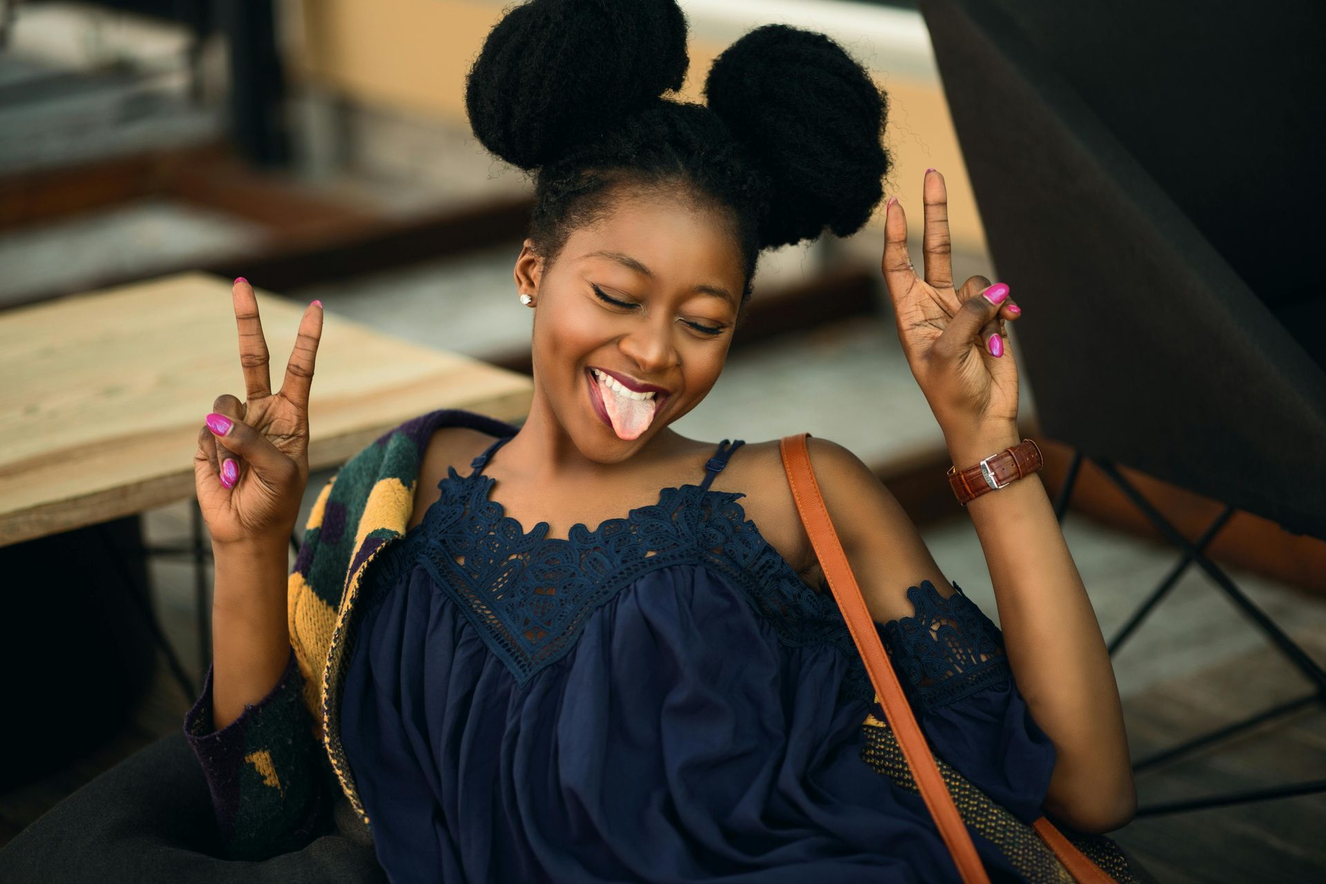 Woman with two space buns making a peace sign and sticking her tongue out. She's in a blue dress, brown bag, and sitting