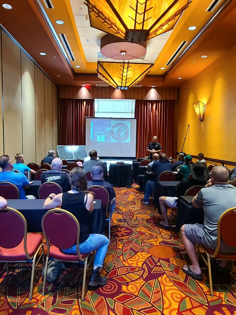 Conference audience seated in a hotel meeting room facing a presentation screen, under warm ceiling lights.