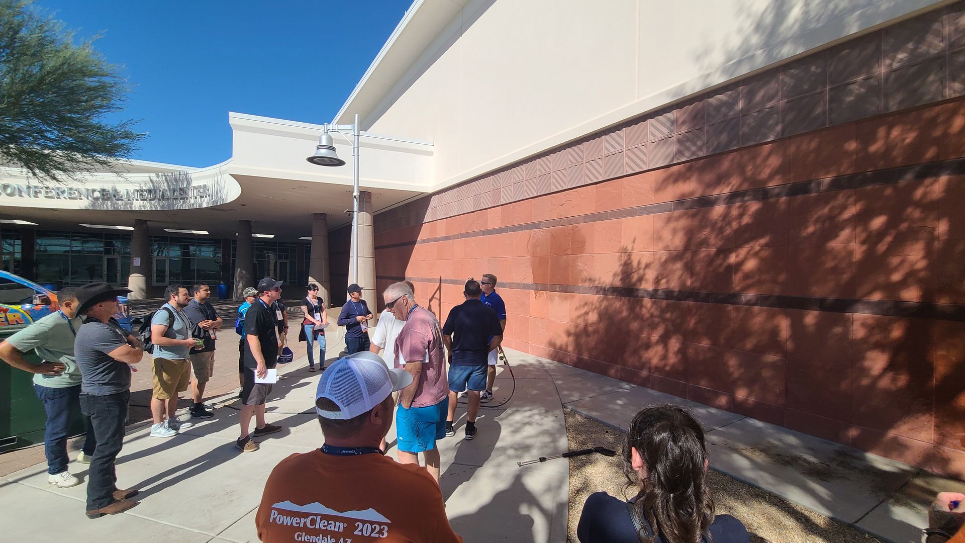 People waiting outside a red brick building in a sunny courtyard, with shadows cast on the wall.
